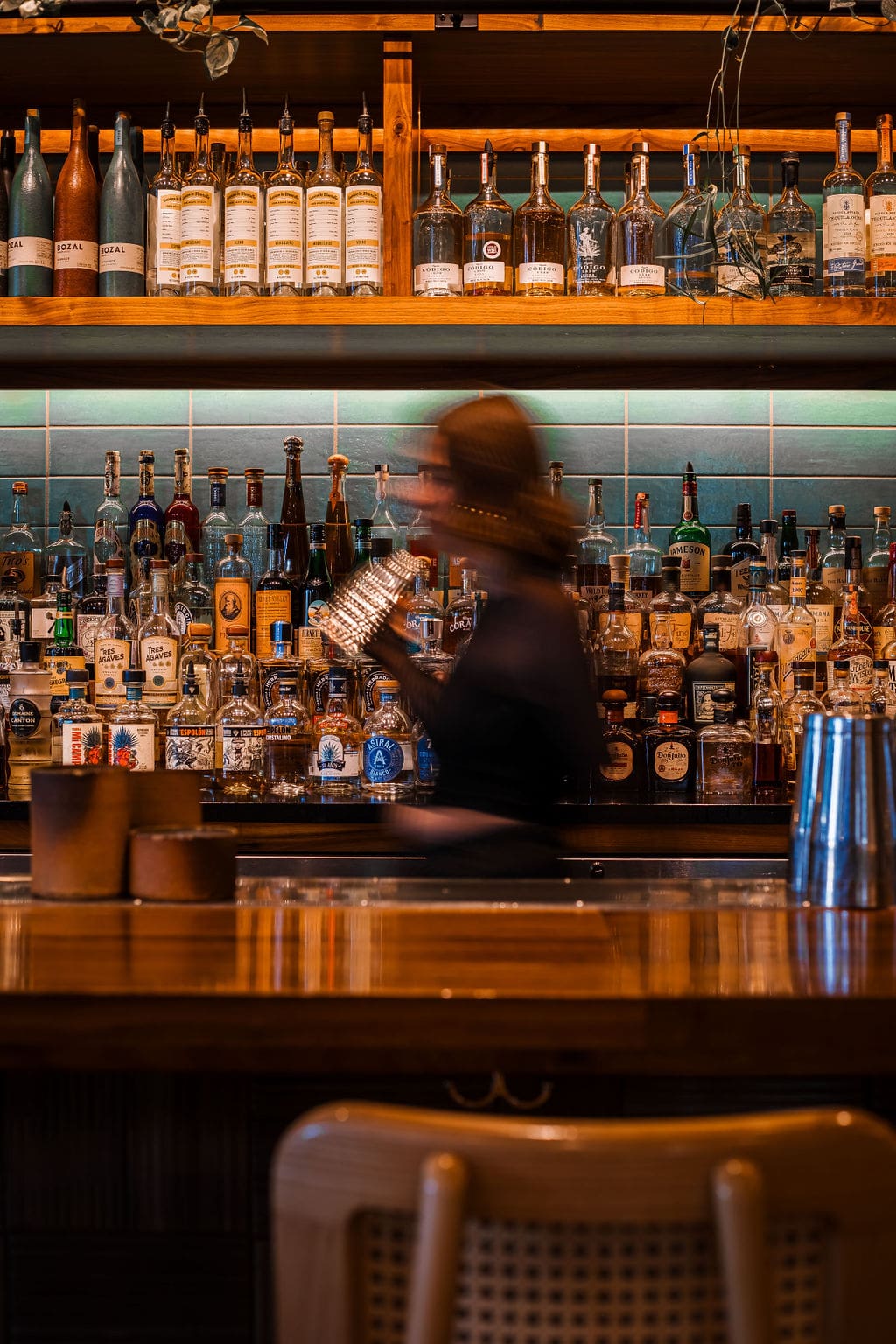 A bartender is blurred as they walk behind a wooden bar top and shake a cocktail