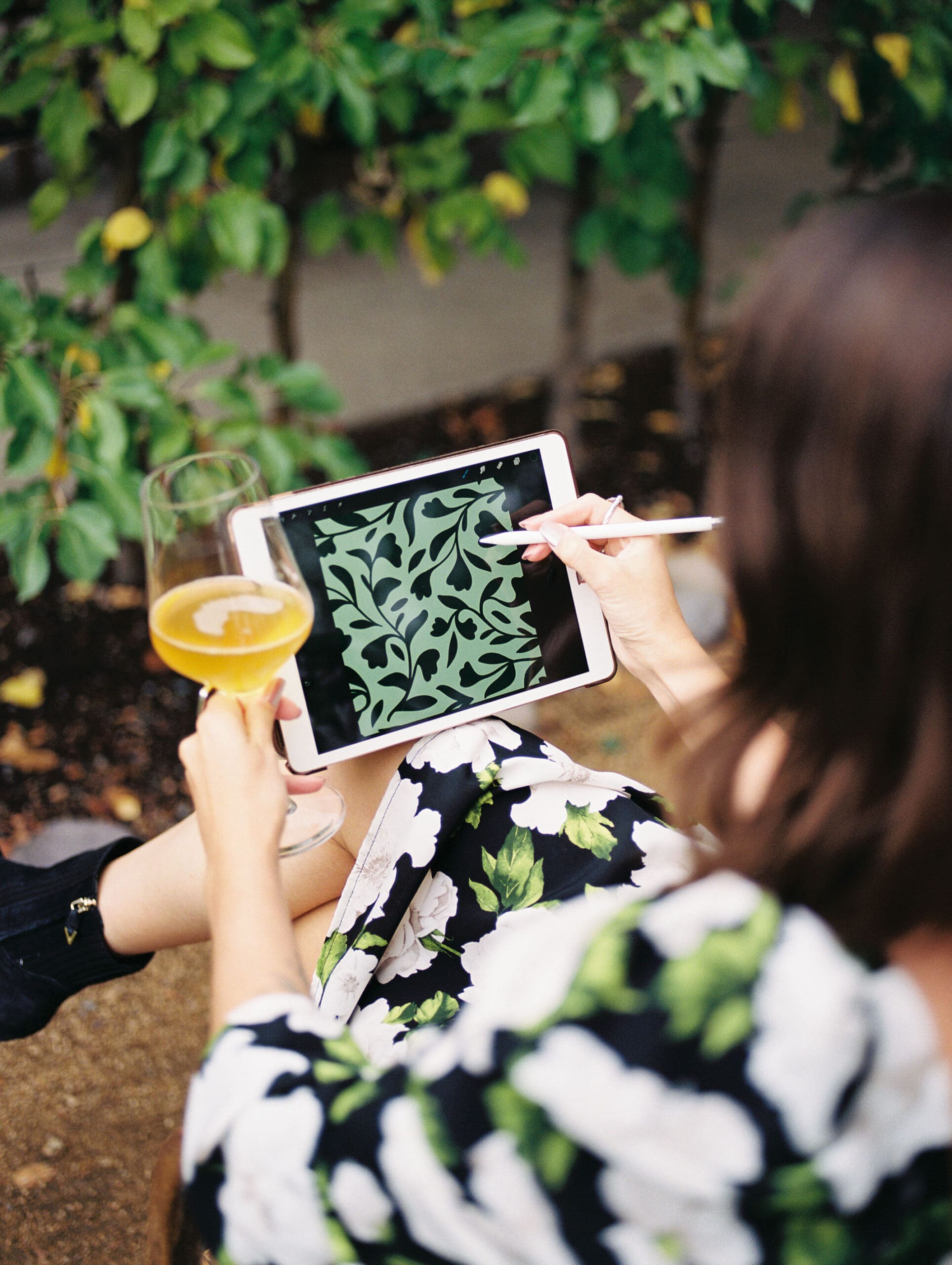 Emma with Bright Spot Studios is wearing a green, white, and black floral dress and sips on an apple cider and draws a green floral pattern on her ipad