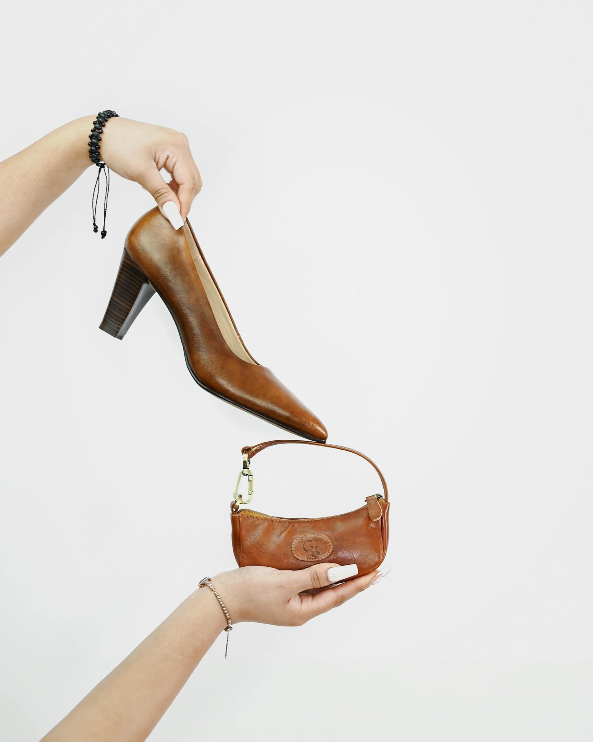 Two arms hold up a shoe and a purse against an off-white background