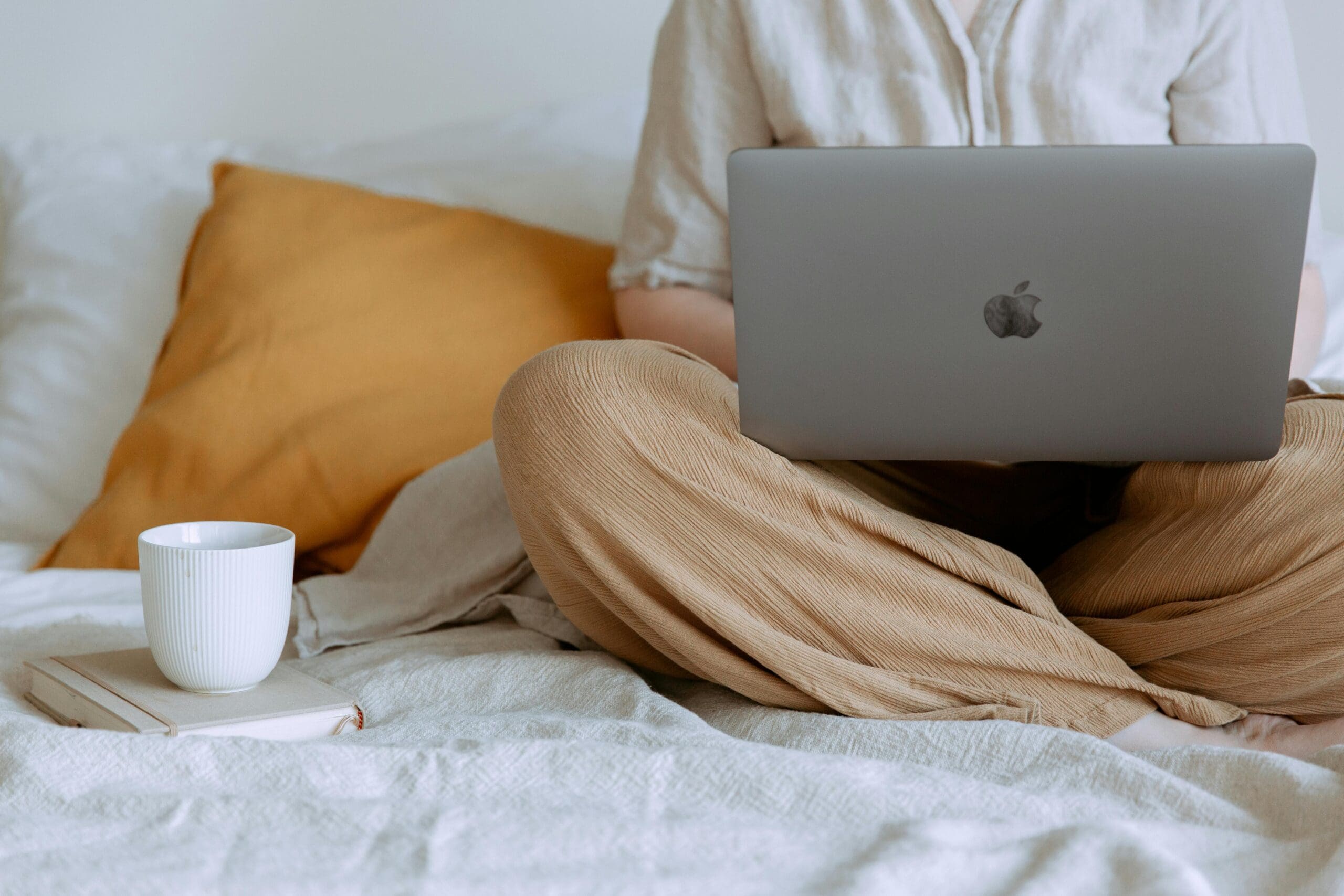 A person sits on a bed using their laptop with a cup of coffee next to them
