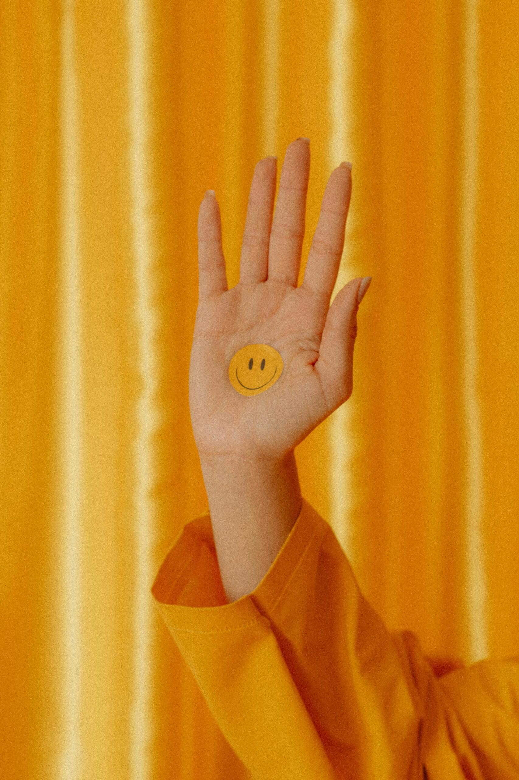 A person holds up the palm of their hand with a yellow smiley face sticker on it against a yellow curtain backdrop